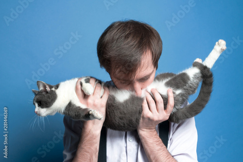 handsome man in shirt and black  suspender holding and kissing stomach cute gray and white cat on blue background © Viktoriia