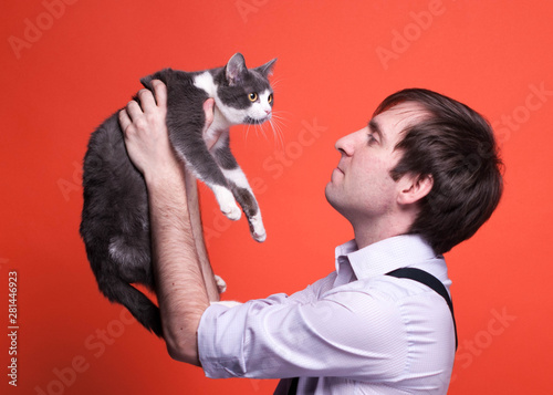 side view of handsome man in shirt with rolled sleeves holdind  in outstrtched hands cute greywith white cat in front of orange background with copy space