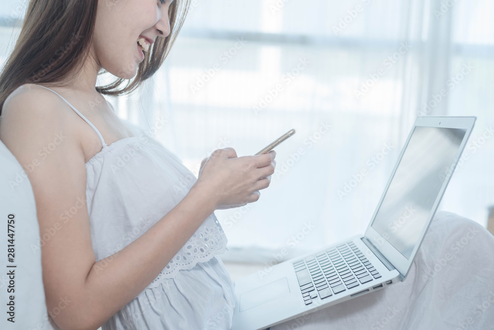 Naklejka premium Happy woman reading news as she relaxes in bed with smartphone and laptop computer in the morning