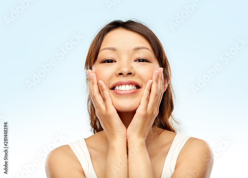 beauty, skincare and people concept - happy smiling young asian woman touching her face over blue background