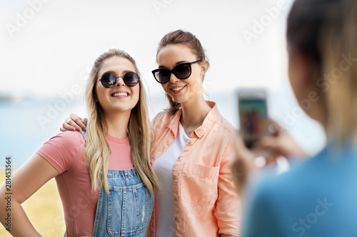 leisure and friendship concept - happy smiling teenage girls or best friends in sunglasses being photographed by smartphone at seaside in summer