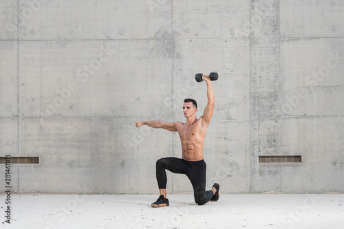 Muscular shirtless caucasian male athlete performs a dumbbell weightlifting exercise in a grungy concrete structure while showing his six pack abs 