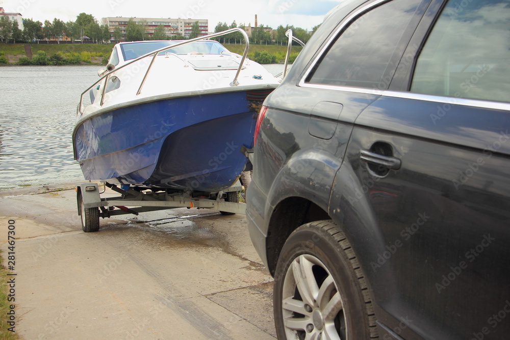 A 4x4 black car pulls blue-white trimaran cabin motor boat on trailer on concrete slipway into the river water, DIY boat launch