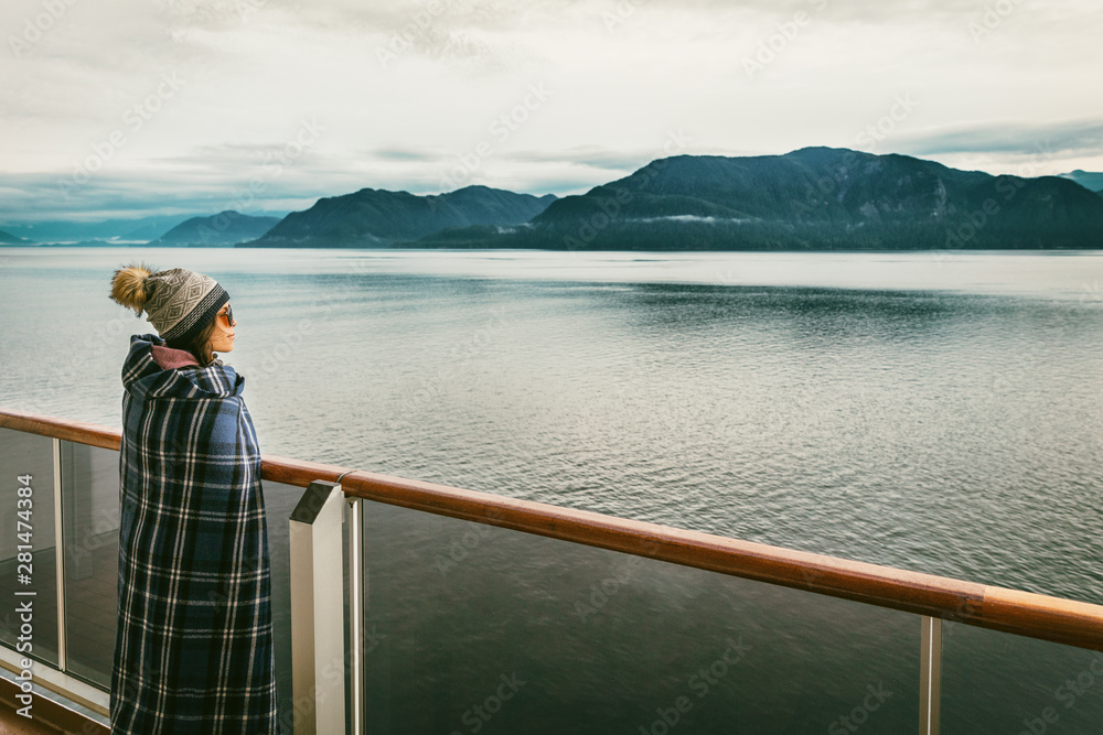 Alaska cruise travel luxury vacation woman watching inside passage scenic cruising day on balcony deck enjoying view of mountains and nature landscape. Asian girl tourist with wool blanket.