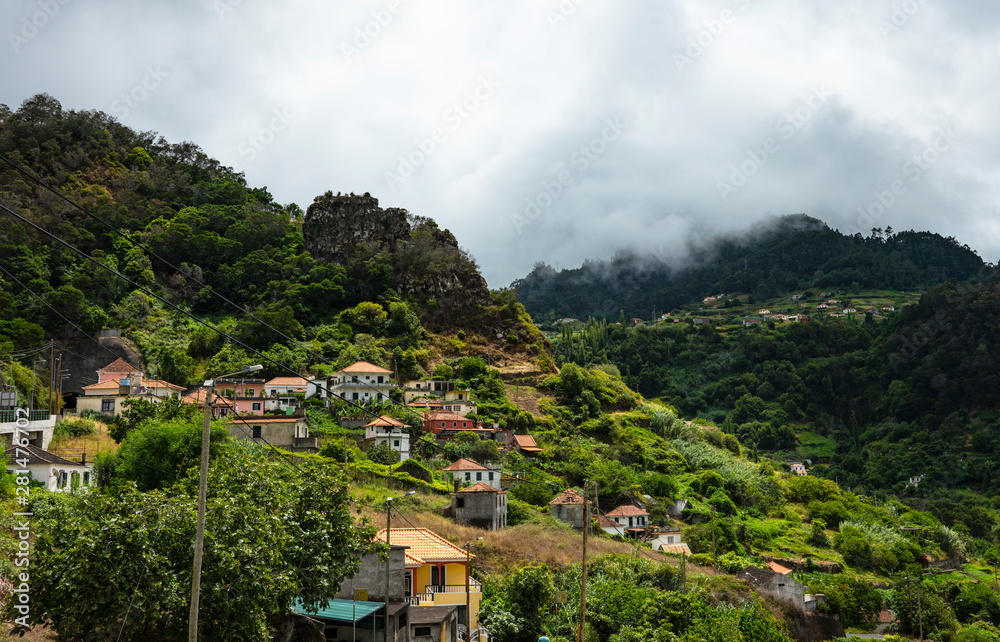 Mountain landscape. A small village is located on the mountainside. Green mountains, small cottages, gardens and orchards are depicted. In the background you can see clouds above the mountain tops.