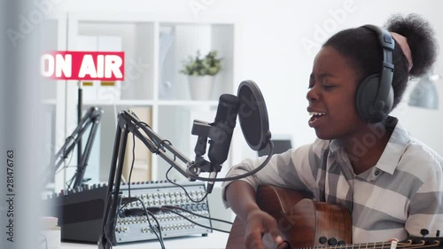 Medium shot of young African woman with headphones on head sitting at microphone in broadcast studio, playing guitar, singing, then looking at camera and smiling photo