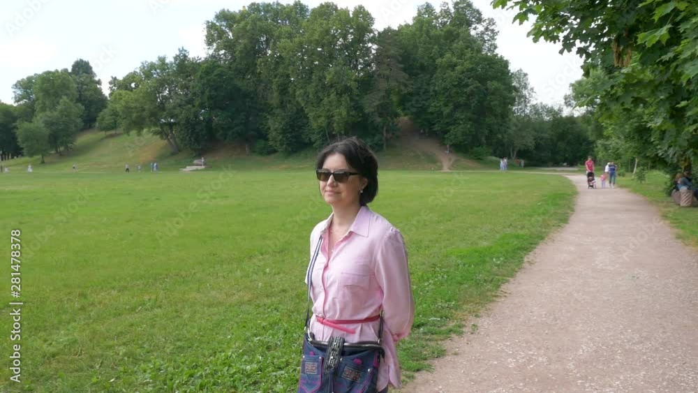 A middle-aged woman in sunglasses, in a pink shirt, with a red belt, with a bag over her shoulder, slowly walks in the park. Along a large meadow, on a sunny summer day. Slow motion A middle-aged woman in sunglasses, in a pink shirt, with a red belt, with a bag over her shoulder, slowly walks in the park. Along a large meadow, on a sunny summer day. Slow motion