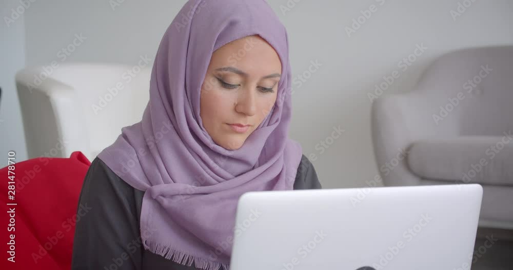 Closeup portrait of young muslim businesswoman in hijab using the laptop sitting in the seatbag looking at camera smiling happily indoors in the apartment Closeup portrait of young muslim businesswoman in hijab using the laptop sitting in the seatbag looking at camera smiling happily indoors in the apartment