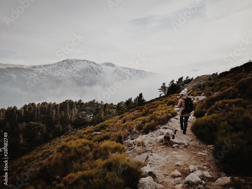A mountaineer is walking with her dog along the mountain path. In the distance you can see the snowy mountains and thick fog.