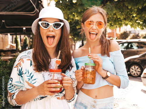 Two young beautiful smiling hipster girls in trendy summer clothes and panama hat.Sexy carefree women posing in the street.Positive models having fun in sunglasses.Drinking fresh cocktail smoozy drink