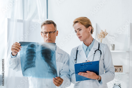 attractive doctor holding clipboard and looking at x-ray near coworker photo