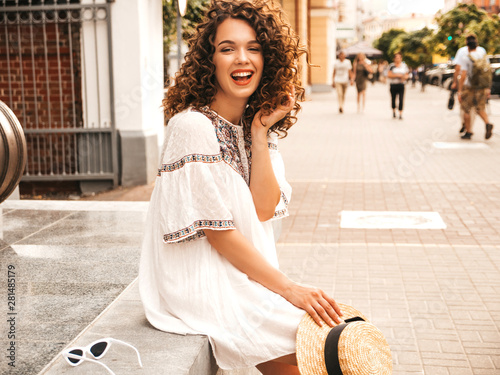 Beautiful smiling model with afro curls hairstyle dressed in summer hipster white dress.Sexy carefree girl posing in street.Trendy funny and positive woman having fun in hat