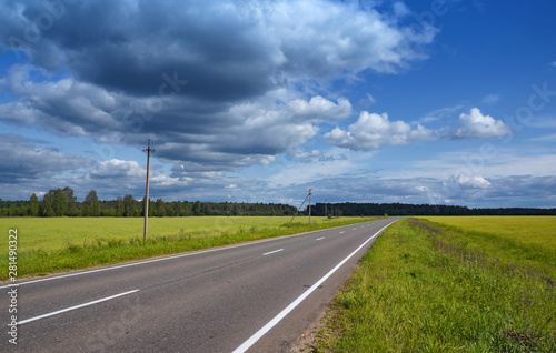 Empty asphalt road in the countryside going through the fields, forest in the background. Sunny summer day, blue sky with clouds. View from right side