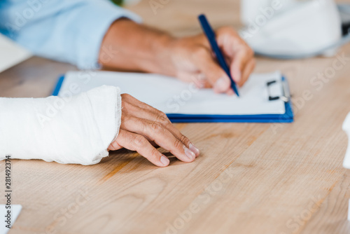 Foto cropped view of injured man with gypsum bandage on broken arm writing on clipboa