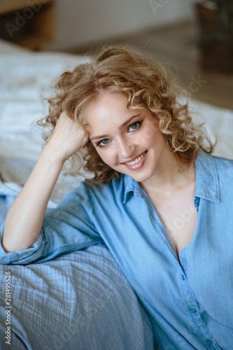 Pretty cheerful blonde young woman with curly hair and charming smile resting in her home, room with modern scandinavian interior