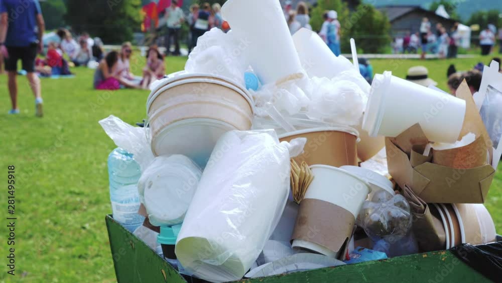 Plastic and paper trash in a crowded container in the park next to the cafe. In the background, people relax on the green lawn at a festival or village fair.