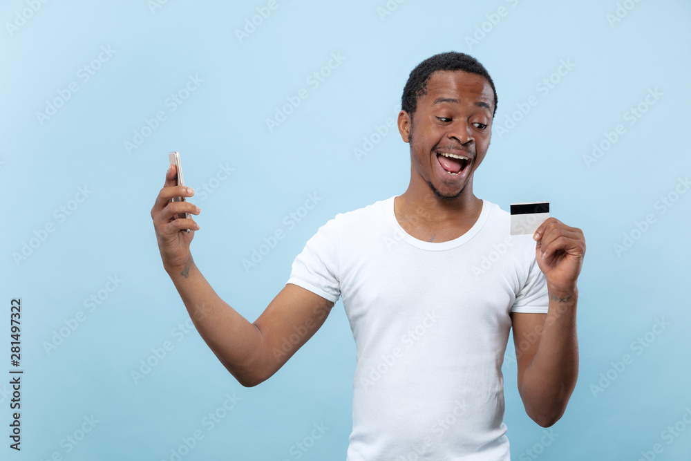 Half-length portrait of young african-american man in white shirt holding a card and smartphone on blue background. Human emotions, facial expression, ad, sales, finance, online payments concept. Half-length portrait of young african-american man in white shirt holding a card and smartphone on blue background. Human emotions, facial expression, ad, sales, finance, online payments concept.