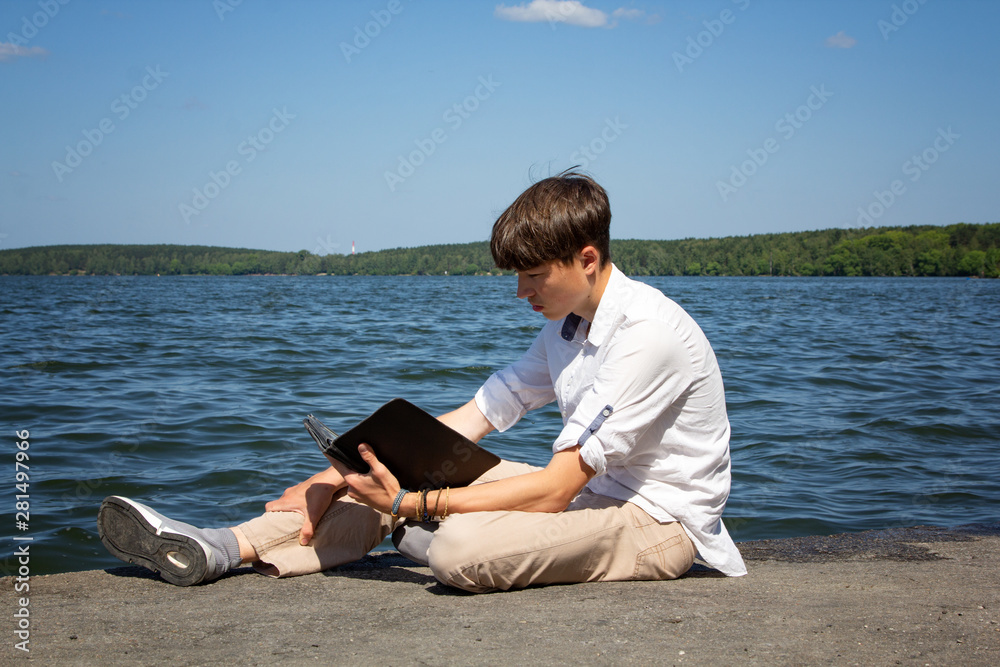 Portrait of a teenager, man, in a white shirt against a blue sky and a large lake. A high school student sits by the lake with a tablet and reads. Young man.