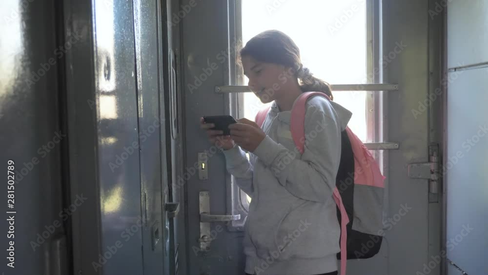 teenager girl traveler with backpack stands by the window of the train car with a smartphone. travel transportation railroad concept lifestyle . the girl in the train at the window corresponds the teenager girl traveler with backpack stands by the window of the train car with a smartphone. travel transportation railroad concept lifestyle . the girl in the train at the window corresponds the