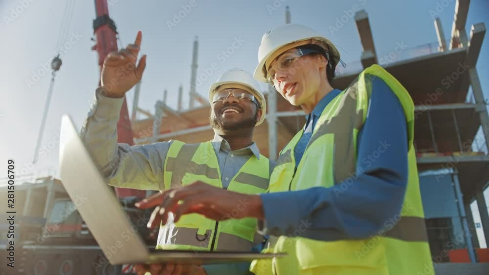 Two Specialists Inspect Commercial, Industrial Building Construction Site. Real Estate Project with Civil Engineer, Investor Use Laptop. In the Background Crane, Skyscraper Concrete Formwork Frames 