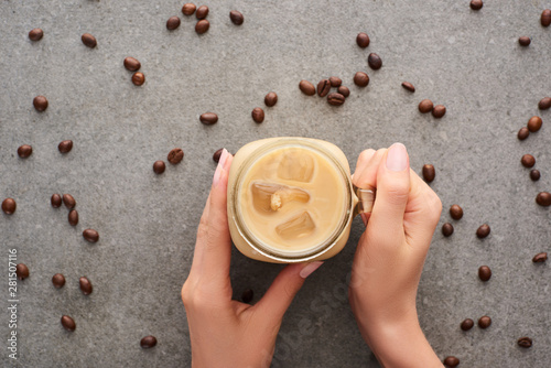 cropped view of woman holding glass jar with ice coffee and coffee grains on grey background
