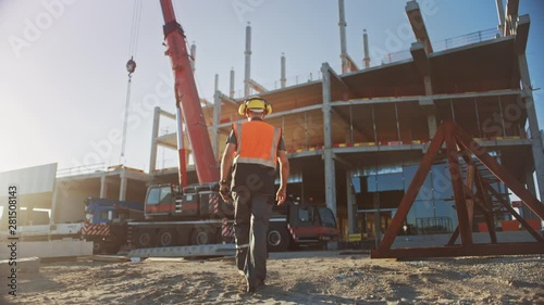 Following Shot: Worker Contractor Wearing Hard Hat and Safety Vests Walks on Industrial Building Construction Site. In the Background Crane, Skyscraper Concrete Formwork Frames 