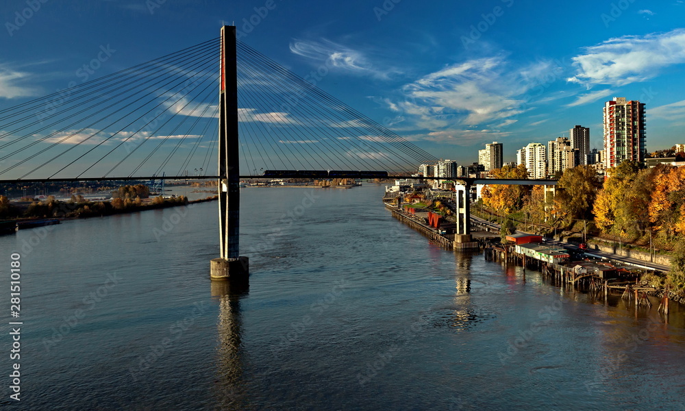 Fototapeta premium New Westminster City the slopes of the Fraser River, painted with autumn colors, Sky Train Bridge and a road against the background of a bright sky with white clouds