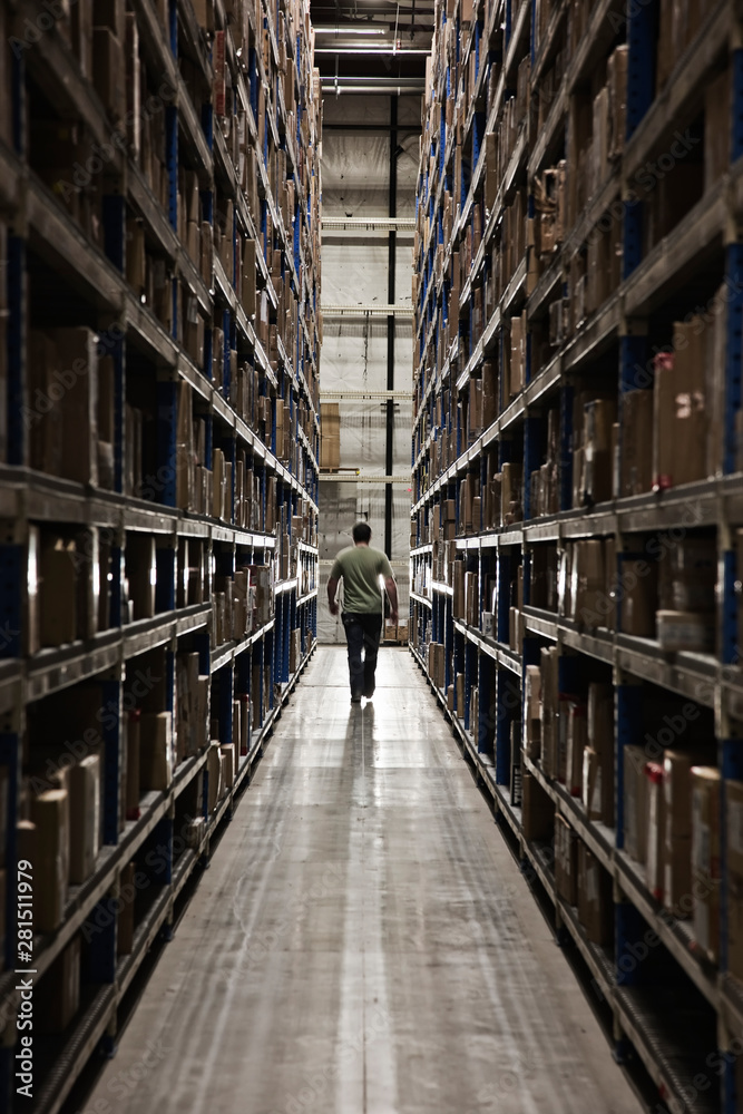 Man walking between racks of cardboard boxes in distribution warehouse Man walking between racks of cardboard boxes in distribution warehouse
