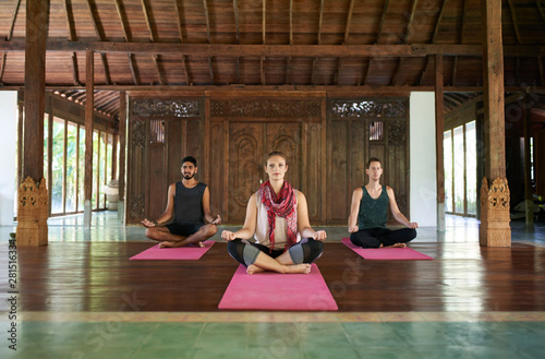 Three multi-ethnic people sitting in lotus pose practicing yoga together in a traditional temple in Bali Indonesia