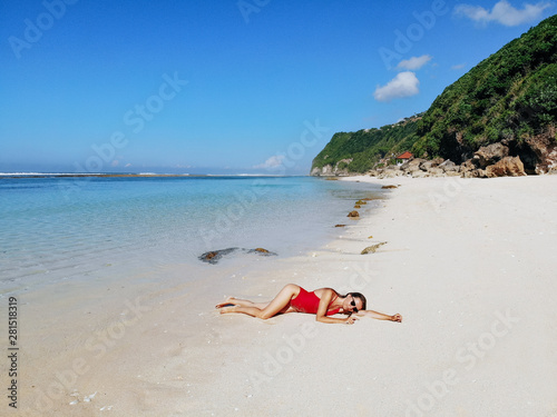 Fototapeta Naklejka Na Ścianę i Meble -  Beautiful woman in red bikini relaxing on white sand tropical beach with blue clear water and jungle. Bali Indonesia. Tropical background and travel concept