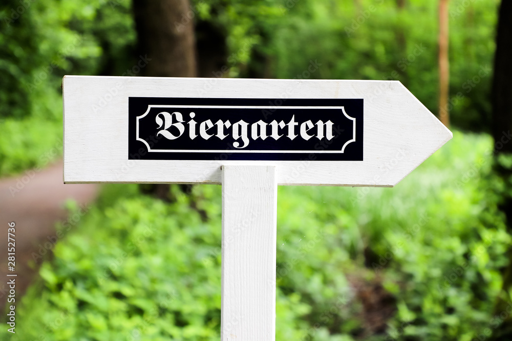Biergarten (Beer Garden in German) white wood arrow direction sign with old German writing style. Sign in open air with green vegetation background outdoors in Germany.