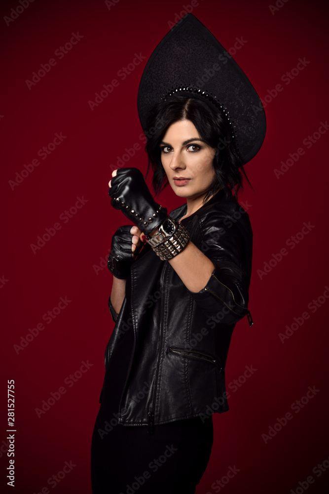 Young stylish brutal woman brunette in neofolk clothes stands in a fighting stance ready to punch on dark red