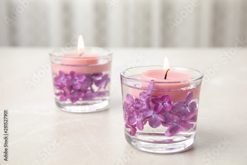 Glass holders with water, lilac flowers and burning candles on light table, space for text