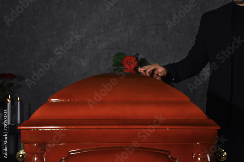 Young man near casket with red rose in funeral home, closeup