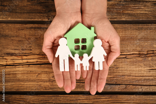 Woman holding figures of family and green house in hands on wooden background, top view photo