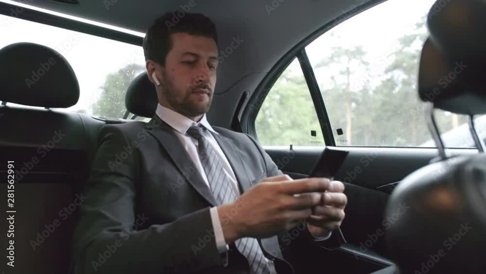 Shaky camera shot of successful businessman in suit and necktie sitting in back seat of moving car and looking at mobile phone Shaky camera shot of successful businessman in suit and necktie sitting in back seat of moving car and looking at mobile phone