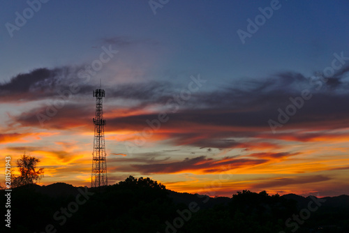 Tall mobile cell phone tower on the high hill sending signal to connect people around the world in the evening when sunset with orange red sky and clouds with copy space on the right.