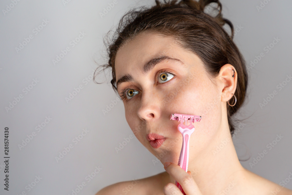 A young woman looks away and tries to shave her cheek with a razor. The concept of getting rid of unwanted hair. Copy space A young woman looks away and tries to shave her cheek with a razor. The concept of getting rid of unwanted hair. Copy space