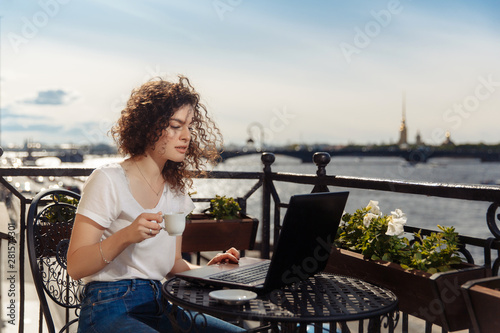 Cute happy young girl working on computer and smiling. Beautiful woman with laptop on a balcony with a landscape on the river Neva and the city of St. ..Petersburg