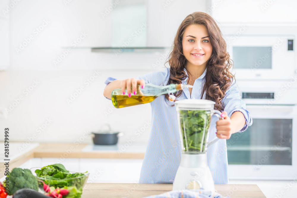 Woman making vegetable soup or smoothies with blender in her kitchen. Young happy woman preparing healthy food or drink with olive oil and fresh vegetable