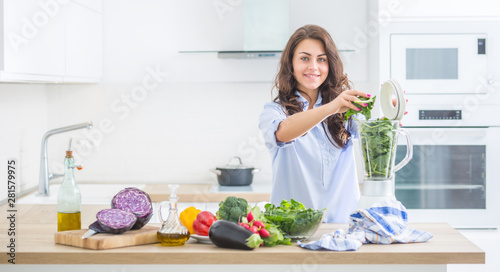Woman making vegetable soup or smoothies with blender in her kitchen. Young happy woman preparing healthy drink with spinach broccoli arugula and other vegetable.