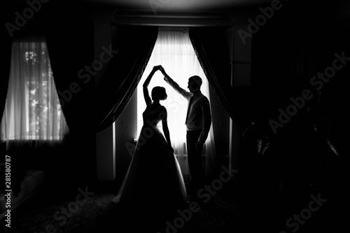 Silhouette of bride and groom by the window. Bride and groom dancing. Newly married couple. Wedding day. The groom holds the bride's hand. The couple dancing the waltz. Wedding dance rehearsal