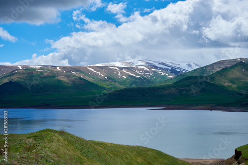 view of a mountain lake against the backdrop of a mountain with snow-capped peaks on a sunny day with clouds in the sky.