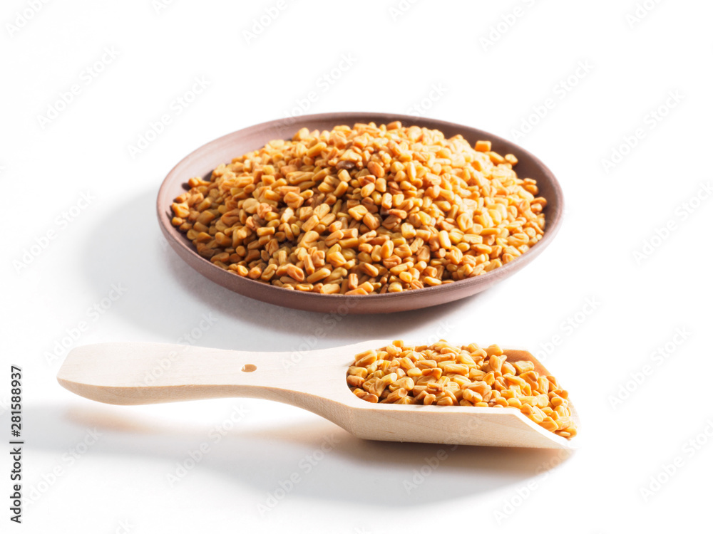 Fenugreek (Trigonetla) on a clay plate with a wooden scoop in the foreground on a white background Fenugreek (Trigonetla) on a clay plate with a wooden scoop in the foreground on a white background