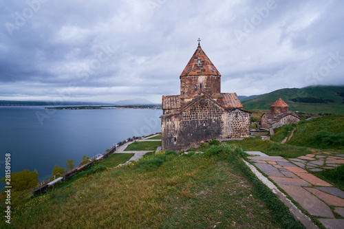 The ancient monastery is located on top of the endless water expanses of the Lake Sevan on an overcast day with clouds in the sky. © StockAleksey