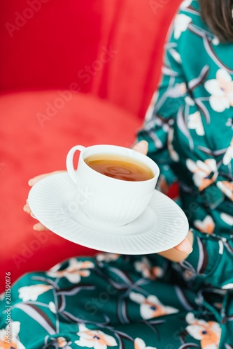 Female hands holding white cup of tea on a red sofa background. Copy space