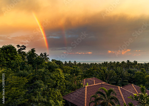 Rainbow over roofs of houses, trees and the sea against the backdrop colourful sunset on a tropical island