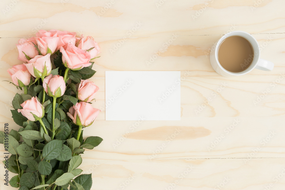 Top view of a white card mockup with a bouquet of pink roses and a coffee on a wooden table.