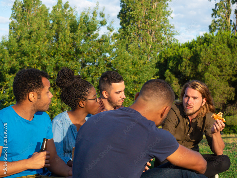 Side view of serious friends having picnic in park. Young people sitting on green grass and eating pizza. Concept of picnic Side view of serious friends having picnic in park. Young people sitting on green grass and eating pizza. Concept of picnic