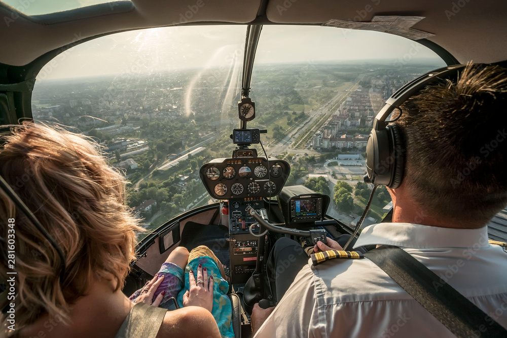 Portrait of beautiful blonde women and pilot enjoying helicopter flight. She is amazed by cityscape. - obrazy, fototapety, plakaty 
